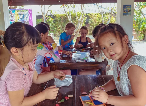 Children making small craft projects together at a shaded outdoor table